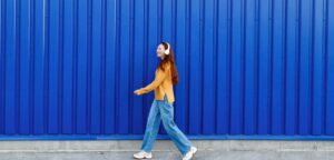 A young woman walks in front of a large blue urban wall wearing white headphones and smiling. Daily walks are a form of rest, which makes you work better.