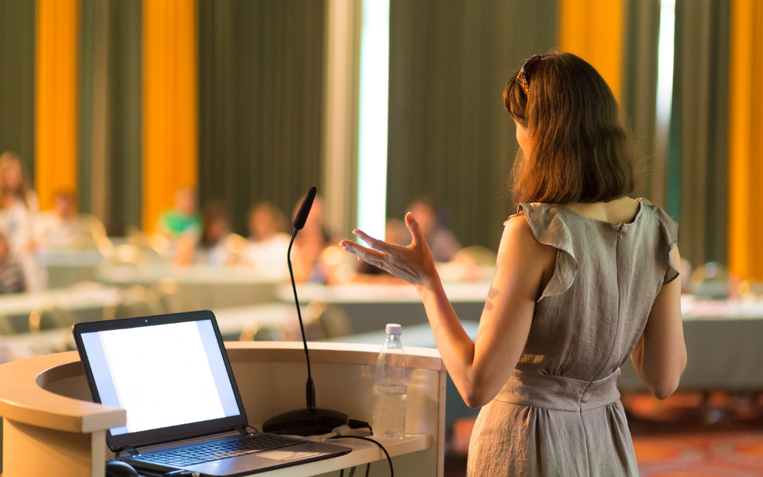 A woman stands at a podium with a laptop, speaking to a group of people seated at tables. Nonprofits can use their Brand for Thought Leadership