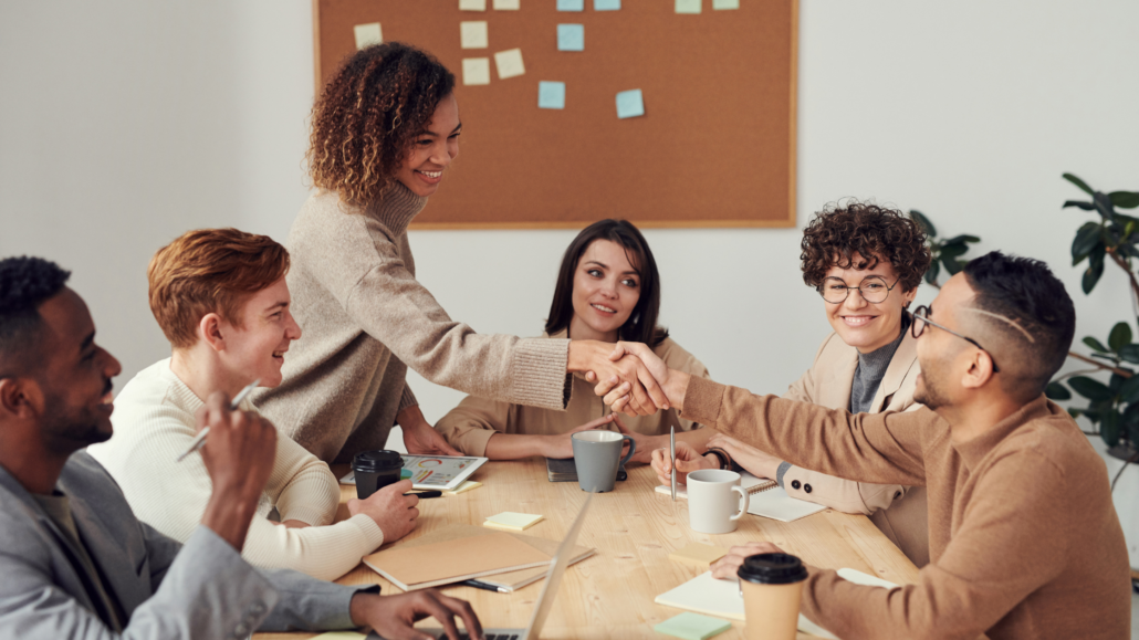 A group of people around a table. They are all smiling and two are shaking hands.